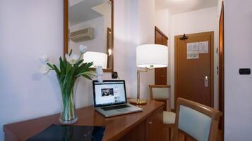 Una habitación de hotel con un escritorio de madera, una computadora portátil, un florero con flores, una lámpara y una silla.