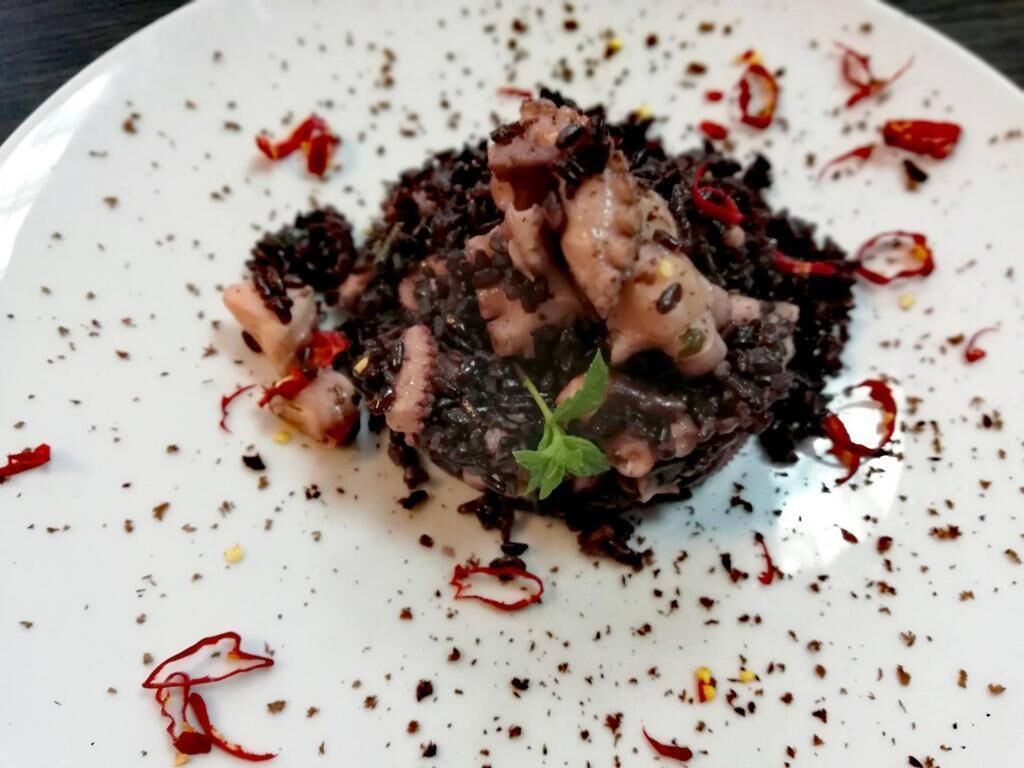A plate with a dish of black rice and meat, garnished with red chili flakes and a green leaf.