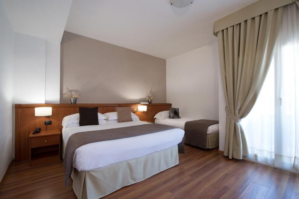 A hotel room with two beds, wooden flooring, a large window with curtains, and a wall-mounted light fixture.