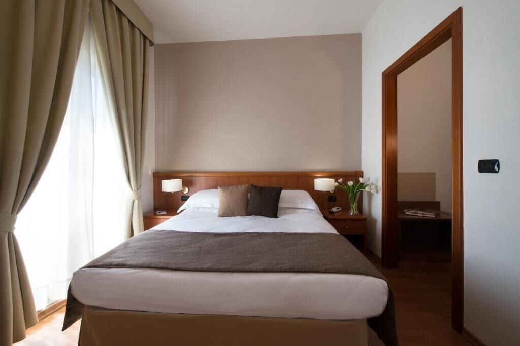 Une chambre d’hôtel avec un grand lit, une tête de lit en bois, des lampes de chevet, une table de chevet et une vue sur un couloir.