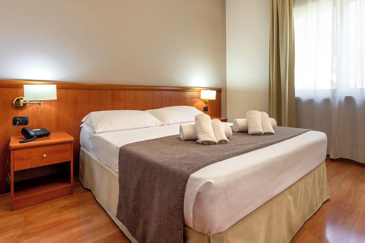 A hotel room with a wooden headboard, a bed with white linens, a brown throw blanket, and two rolled towels on the bed. There is a nightstand with a phone and a lamp.