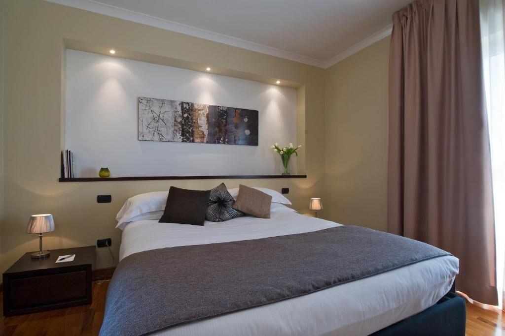 A modern hotel room with a large bed, a bedside table with a lamp, a wall-mounted artwork, and a window with curtains.