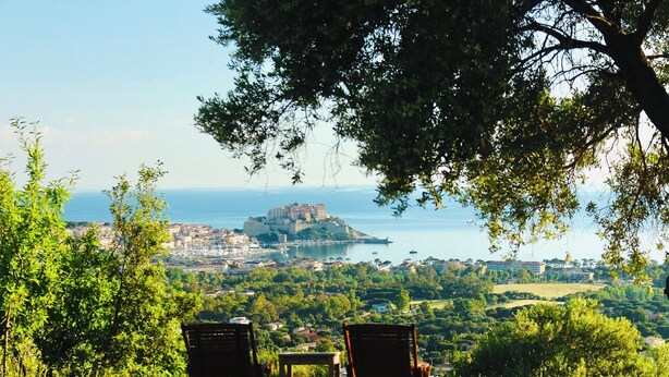 Vista panorámica de un pueblo costero con una estructura imponente al estilo de castillo sobre una colina, que domina un puerto deportivo con barcos. En primer plano, hay dos sillas de madera y una mesita.