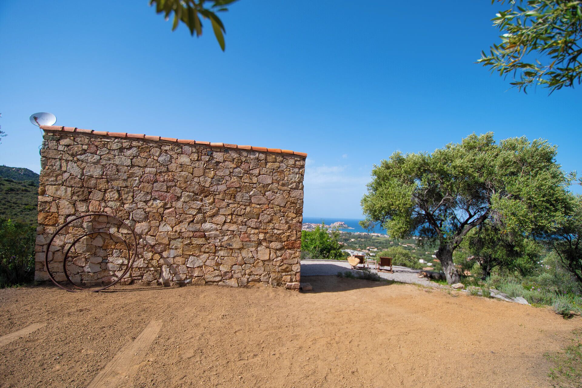 A stone building with a red-tiled roof, a metal gate, and a dirt path leading to a scenic view.