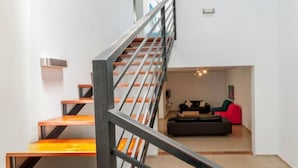 Een trap met metalen leuningen en houten treden die leidt naar een woonruimte met een bank en een rode fauteuil.