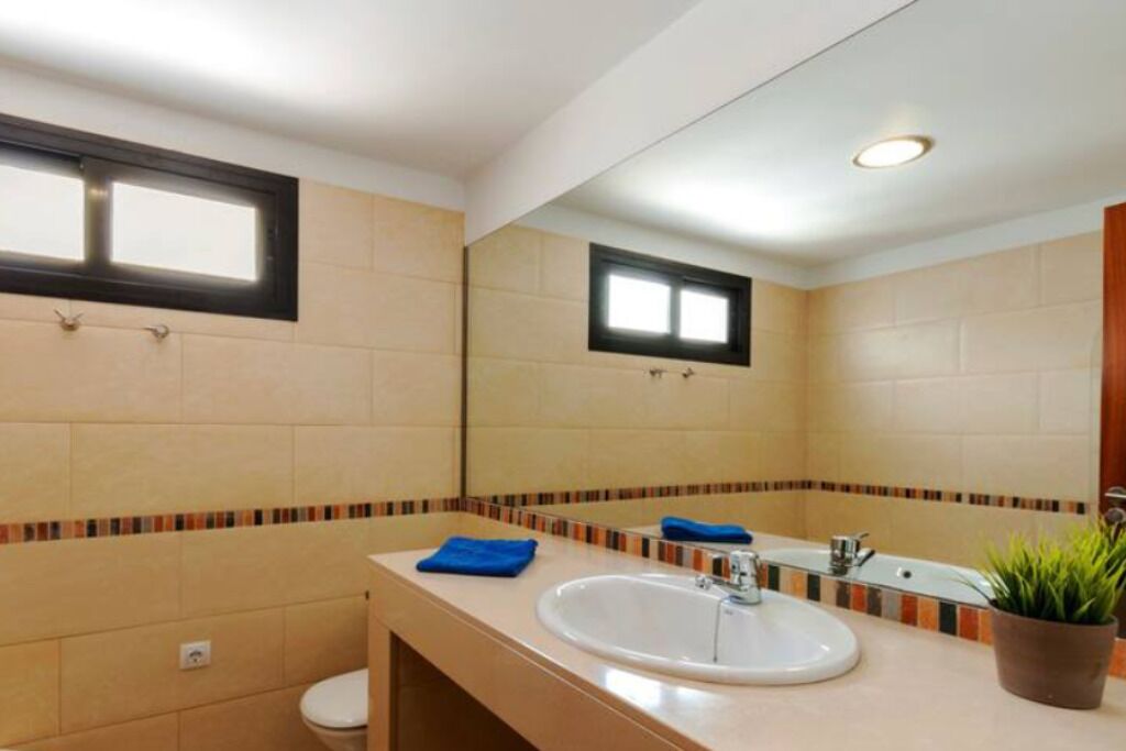 A bathroom with a white sink, a mirror, and a toilet. There is a window above the sink and a small window above the toilet.