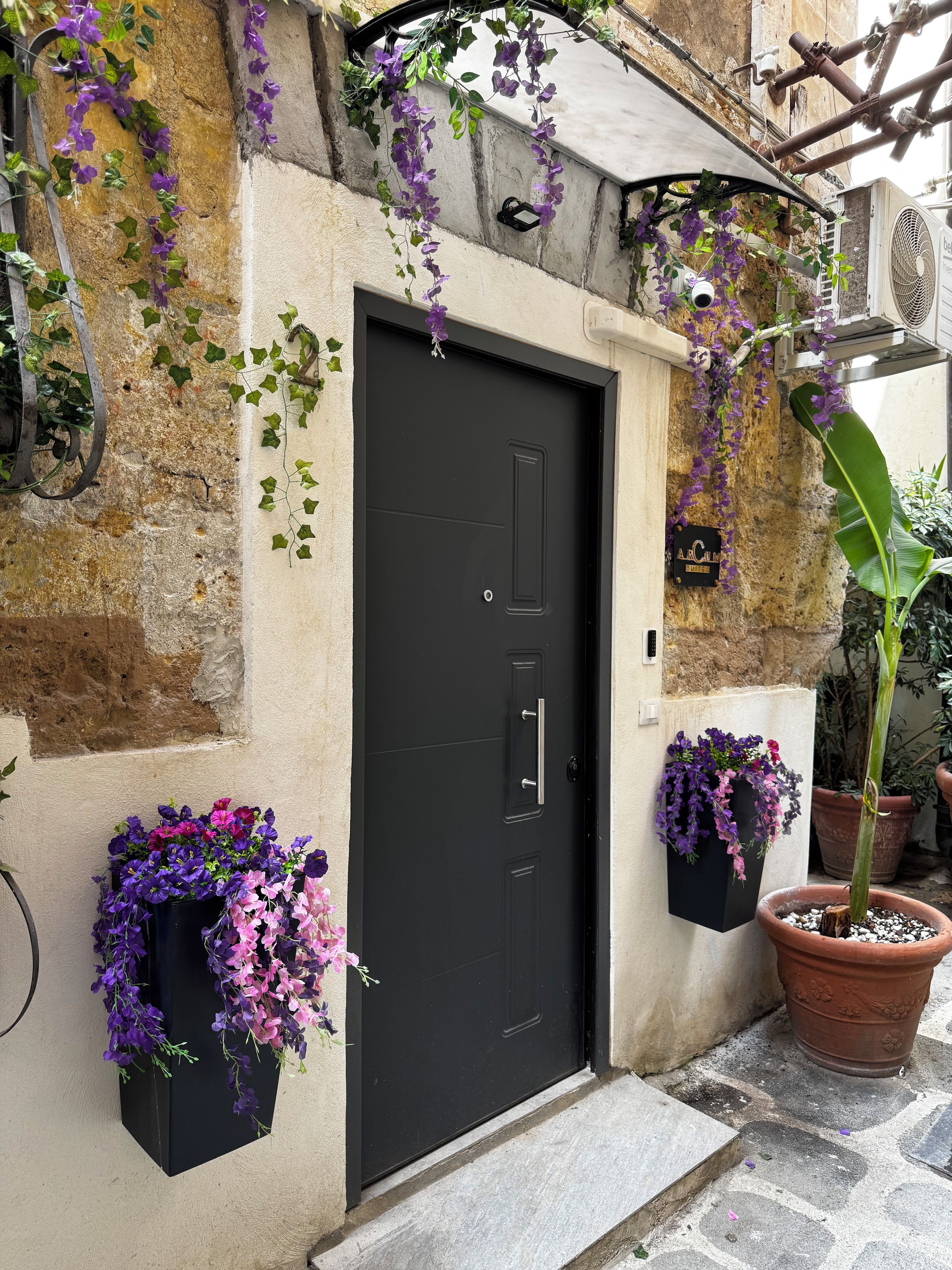 Eine schwarze Tür mit kleinem Fenster, umgeben von Steinmauern und Blumentöpfen mit violetten Blüten.