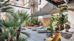 Un patio al aire libre iluminado por el sol con sillones de descanso, plantas en macetas y un muro de piedra.