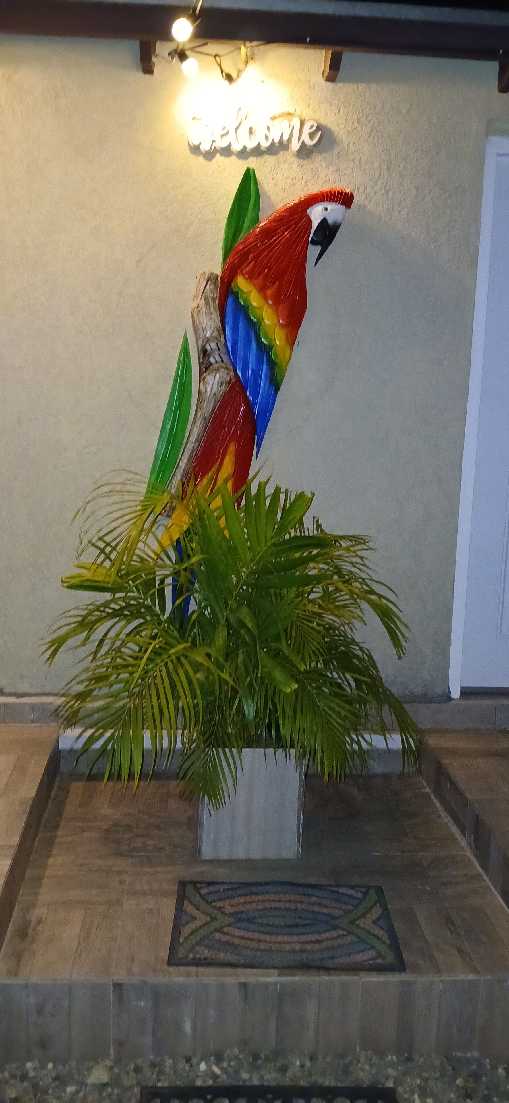 Eine farbenfrohe Papagei-Skulptur mit einem 'Welcome'-Schild darüber, platziert auf einer Holzplattform mit einem gemusterten Teppich davor.