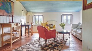 Un salon chaleureux avec un fauteuil rouge, un fauteuil jaune et un canapé blanc. On y trouve une table en bois avec une lampe, une bibliothèque et un tapis à motifs.