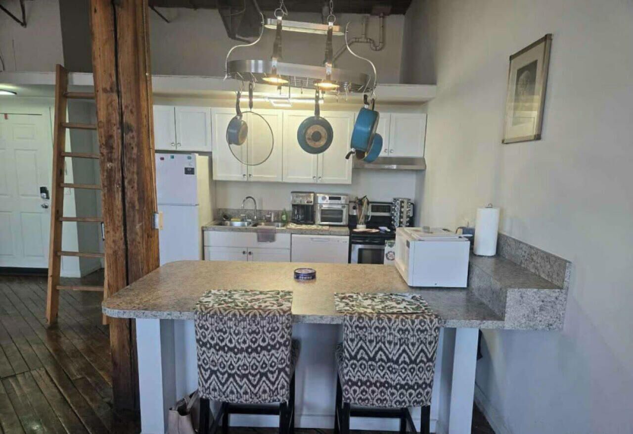 Een keuken met witte kastjes, een centraal eiland met twee gestreepte krukken, hangende pannen en een ladder.