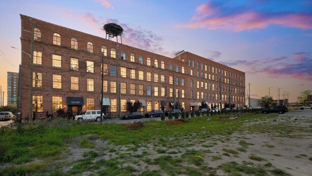 A large brick building with multiple windows and a flat roof, surrounded by a grassy area with parked cars. - Spacious Loft w/ Riverfront Views & Comfort! (Detroit)