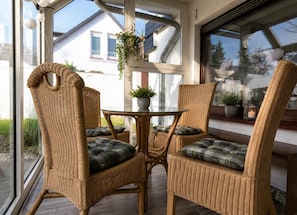 Ein Set aus Korbstuhl und Tisch in einem Wintergarten mit großen Fenstern, Topfpflanzen und Blick auf ein Haus.