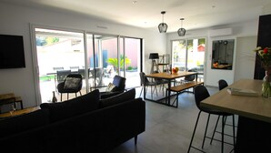 A modern living room with a sofa, a dining table with chairs, and a kitchen area with a bench. - Villa Agapanthes, perfect in any season, with heated swimming pool (Saint-Vincent-sur-Jard)