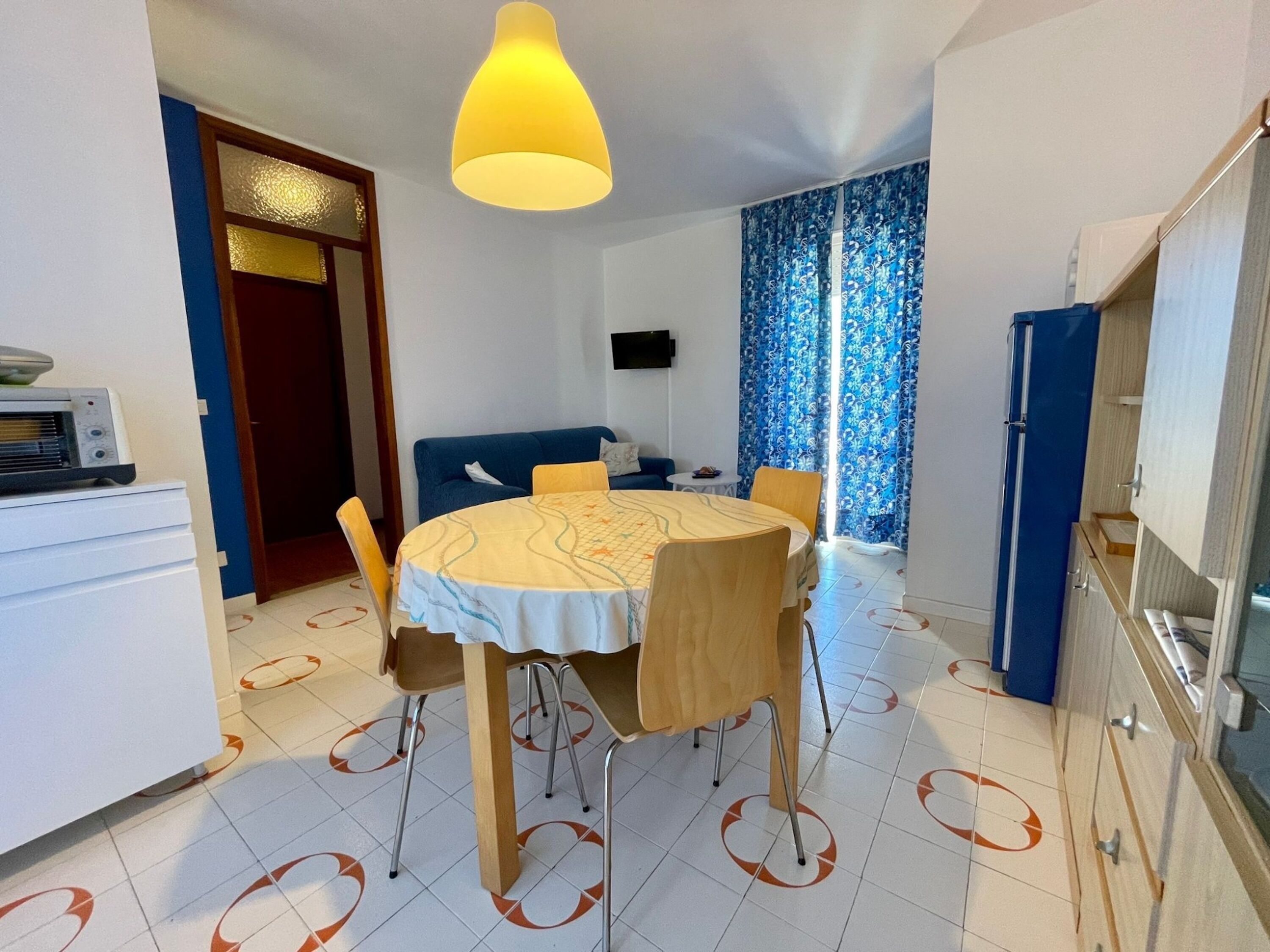 A dining area with a round table, chairs, a yellow pendant light, a blue sofa, a television, and a kitchen area with cabinets and a refrigerator.