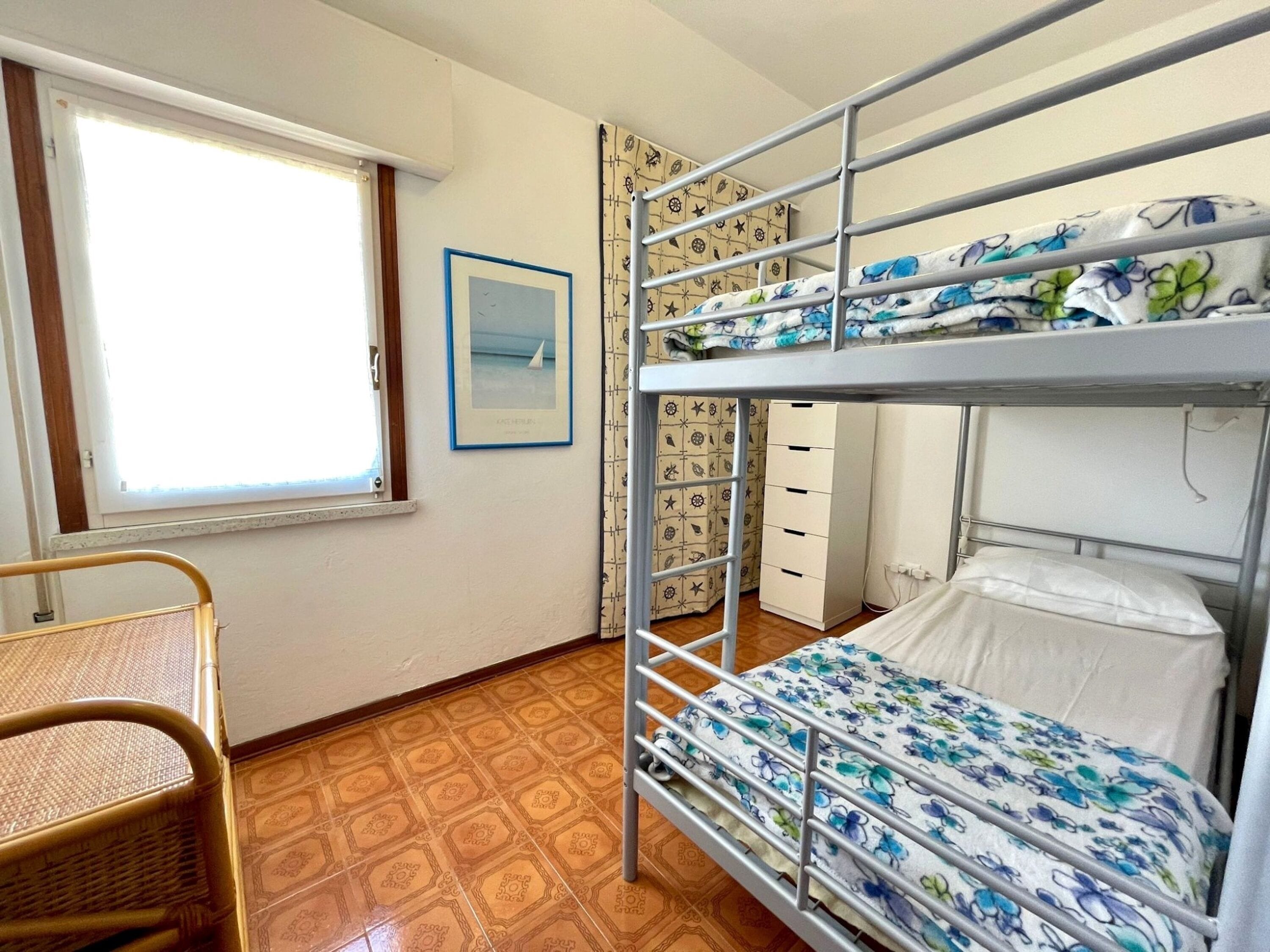 A bunk bed room with a window, a framed picture, patterned wallpaper, and a wicker chair.