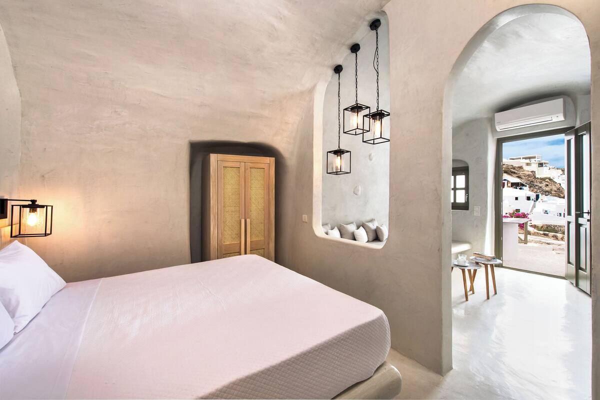 A bedroom with a bed, a wooden door, a window with a view, and a hanging light fixture.