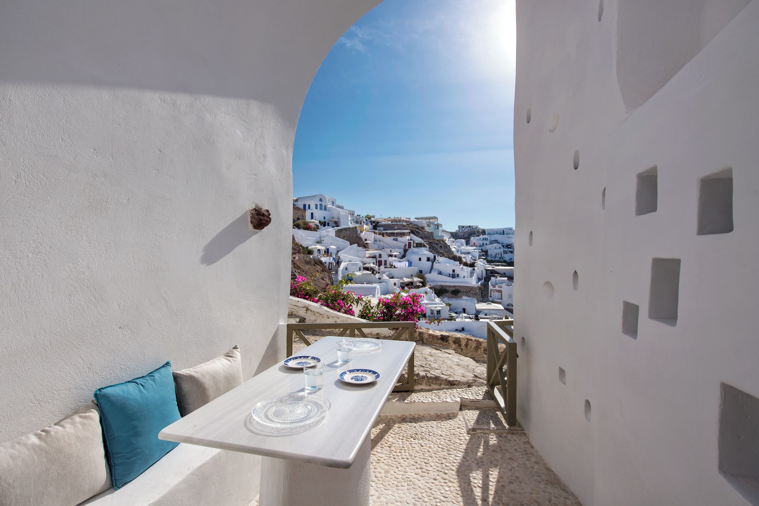 Una mesa blanca con platos y vasos, un almohadón azul, y una vista de edificios blancos y flores rosadas bajo un cielo despejado.