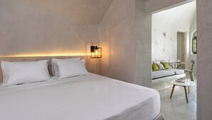 A modern hotel room with a large bed, a bedside table, a wall-mounted light, and a view into a living area with a sofa and a round table. - Rhenia Suites-Prive Jacuzzi&Caldera View (Santorini)