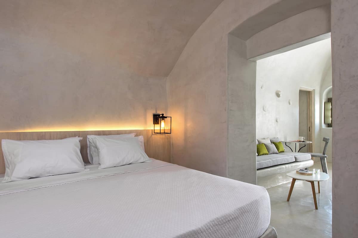 A modern hotel room with a large bed, a bedside table, a wall-mounted light, and a view into a living area with a sofa and a round table.