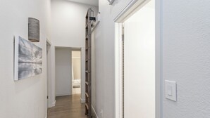 A narrow hallway with a wooden floor, a metal ladder, a wall-mounted light fixture, and a door at the end.