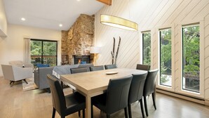A modern dining area with a large table and chairs, a stone accent wall, and large windows offering a view of greenery.