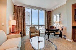 Quarto de hotel com uma janela grande, um sofá, uma mesa de centro de vidro, uma mesa de jantar com cadeiras e um abajur.