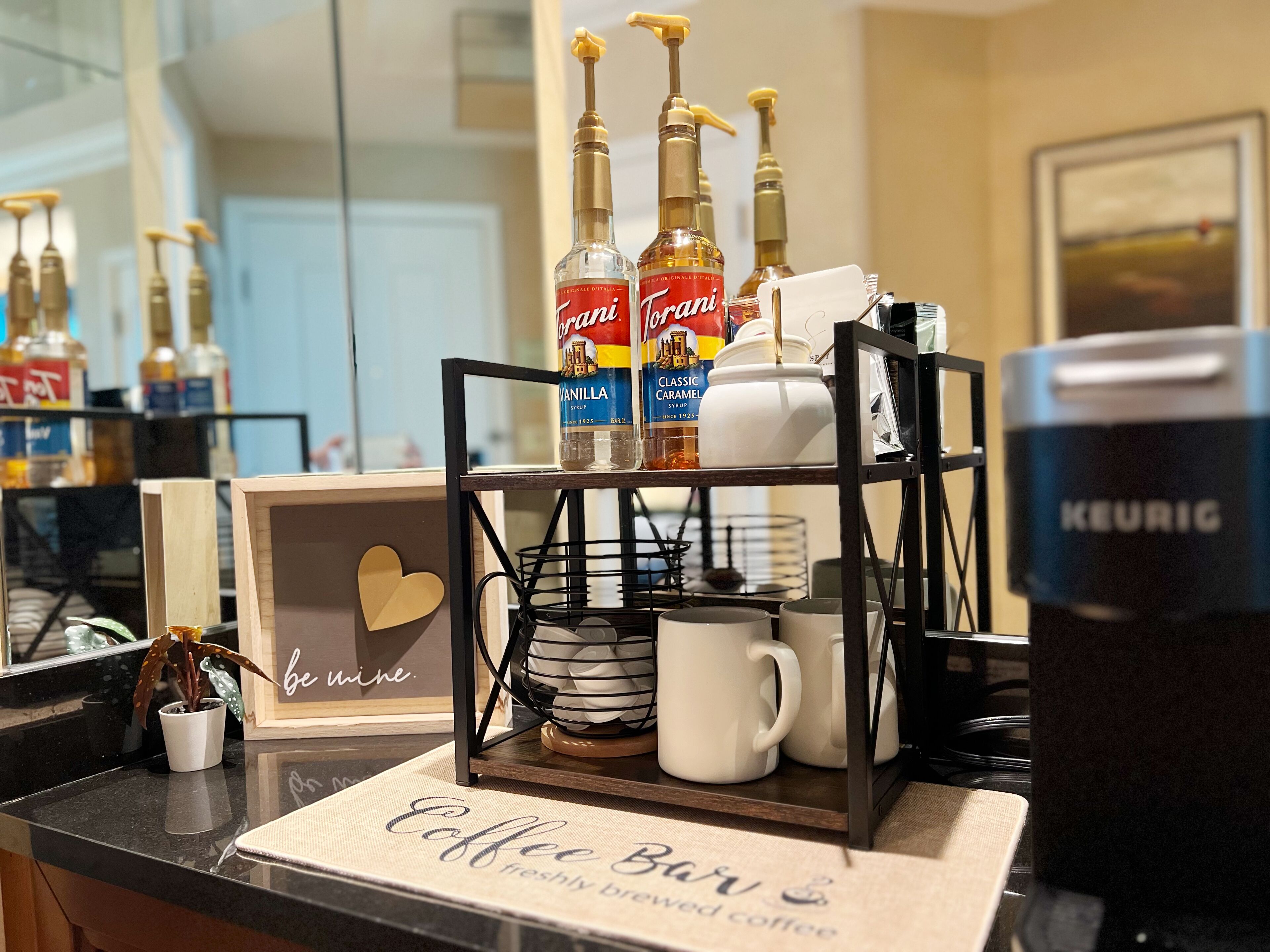 A coffee bar setup with a tray holding Torani syrup bottles, a Keurig coffee maker, and a sign that reads "Coffee Bar freshly brewed coffee."