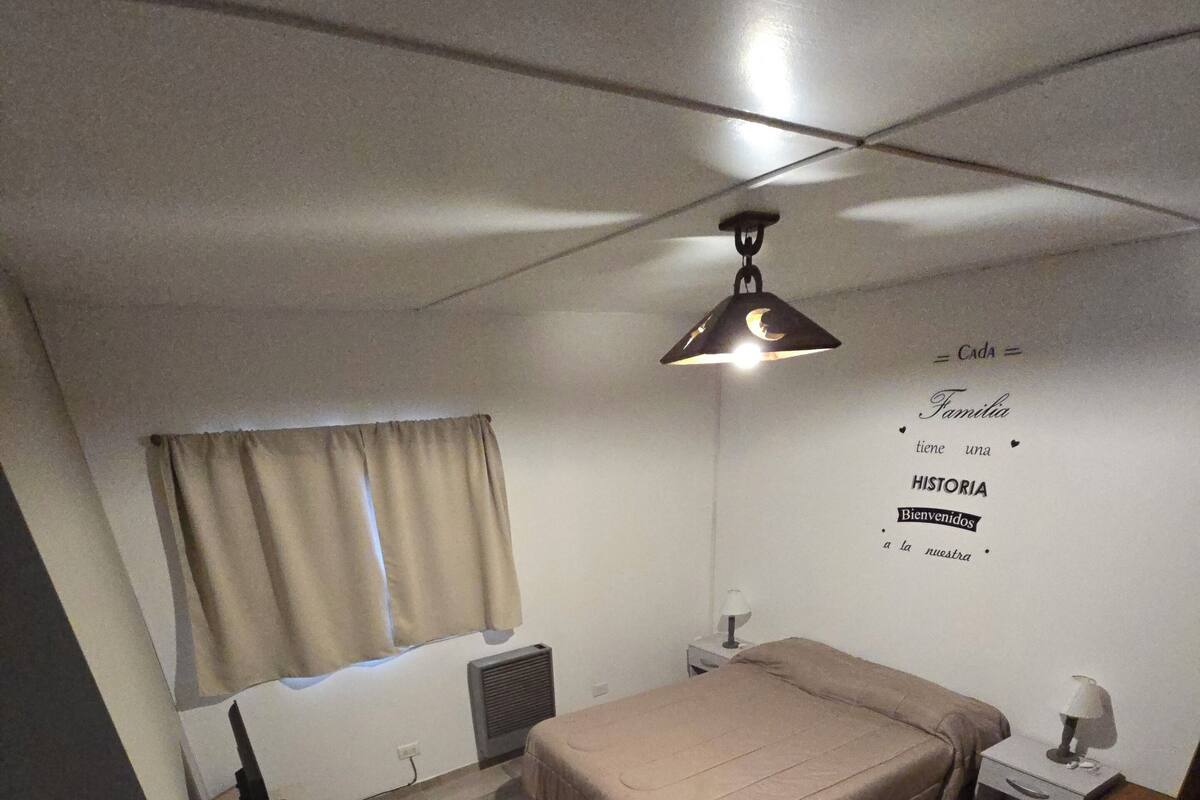Quarto com cama, mesa de cabeceira, abajur, radiador, janela com cortinas e parede com mensagem de boas-vindas.