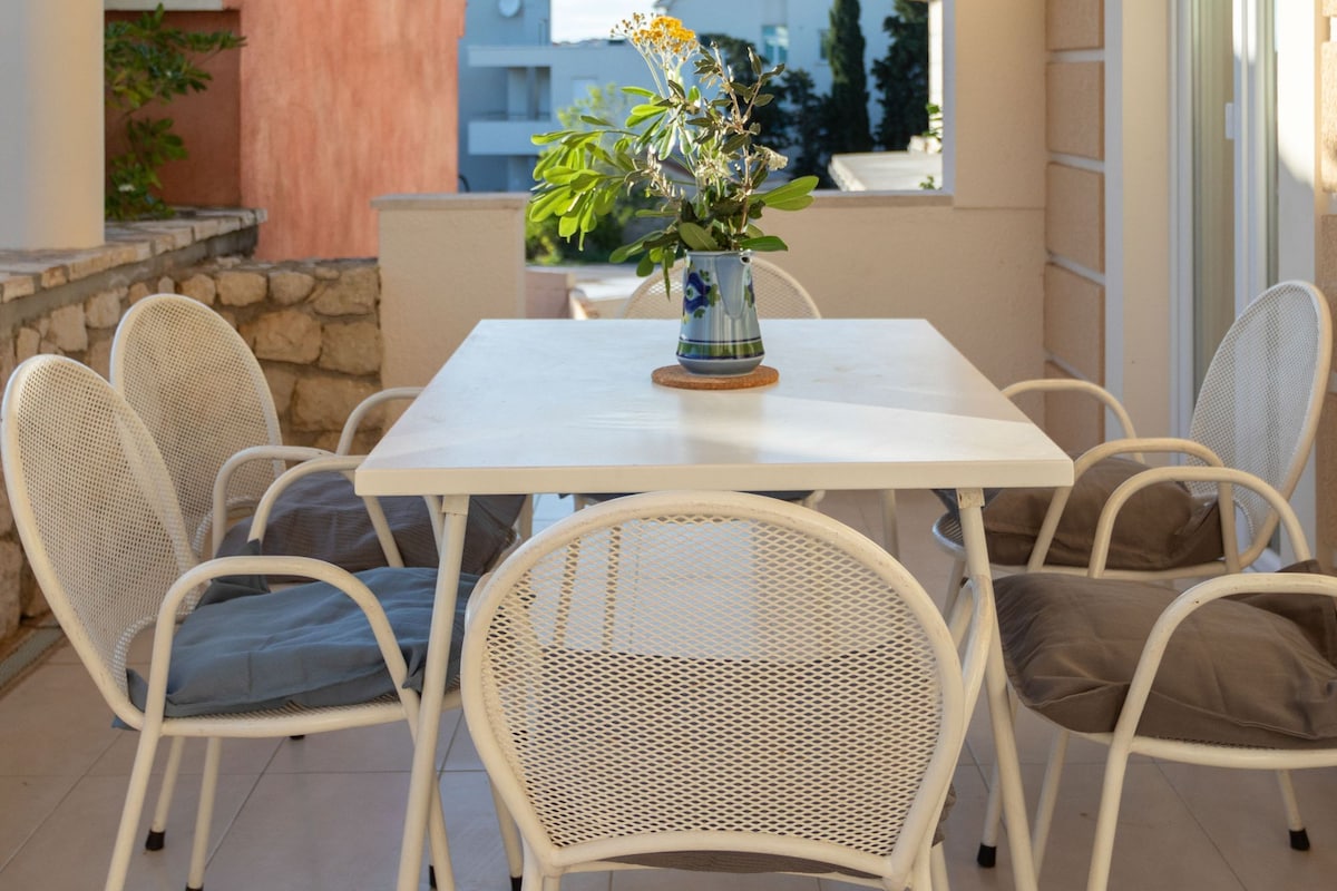 A balcony with a white table and chairs, a vase with flowers, and a view of buildings in the background.