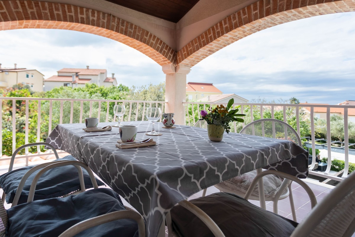 A terrace with a table set for two, surrounded by white metal chairs, a potted plant, and a view of houses in the distance.