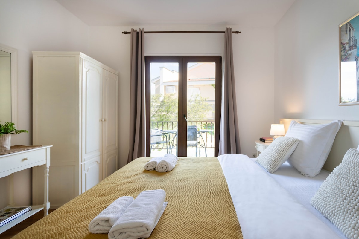 A hotel room with a bed, a desk, a wardrobe, and a balcony with a view of buildings and trees.