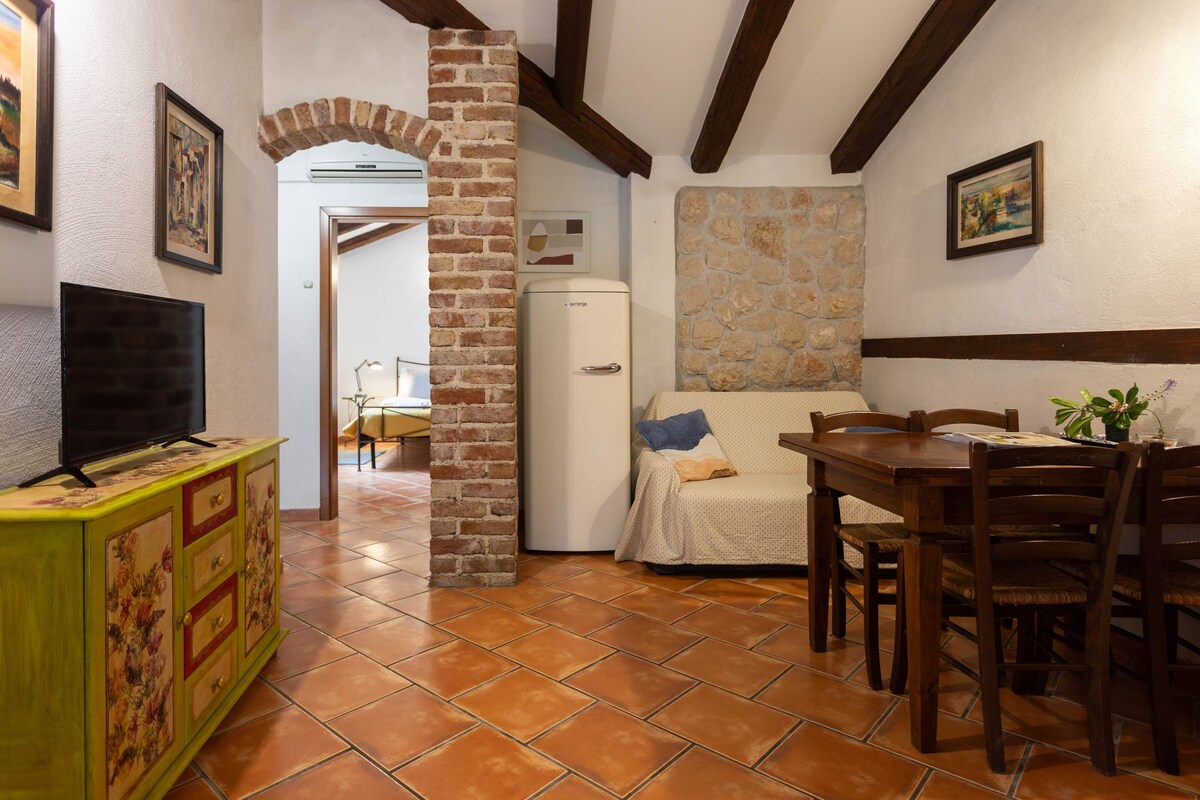 A compact living space with a tiled floor, a dining table with chairs, a sofa with cushions, a television on a cabinet, and a refrigerator.