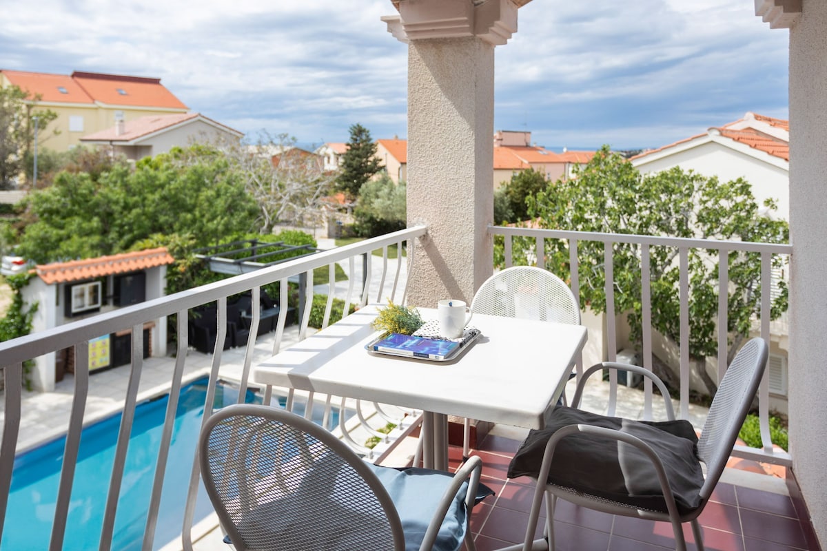 A balcony with a pool, white furniture, and a view of the neighborhood.