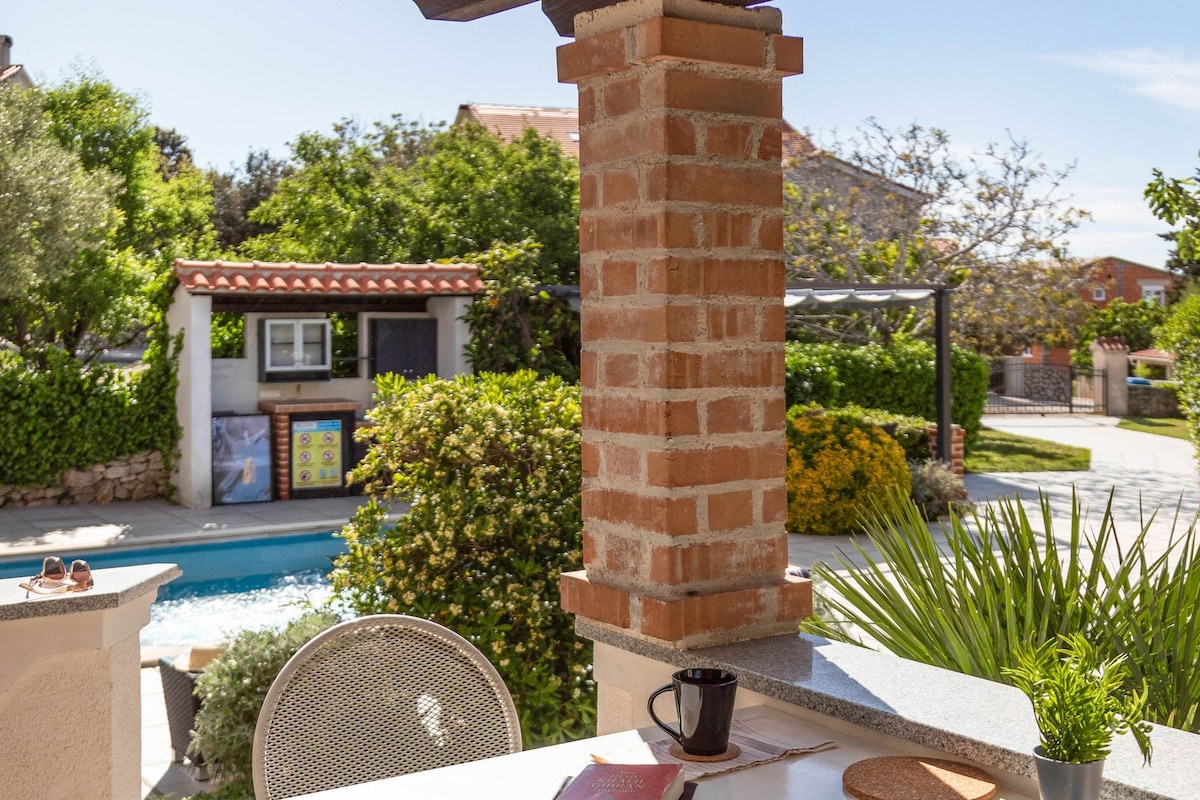 An outdoor patio with a white table, two wicker chairs, a brick pillar, a small potted plant, a pair of sunglasses, a book, and a mug.