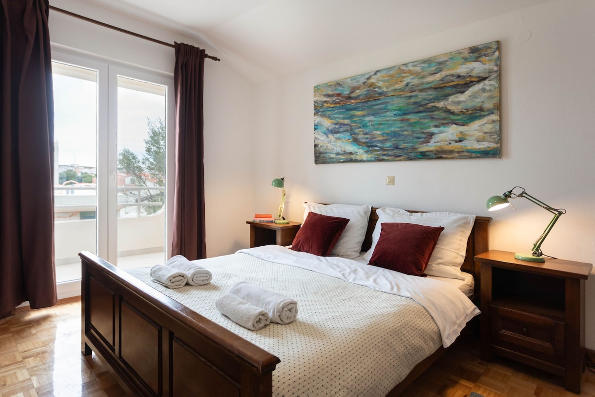 A hotel room with a bed, a painting on the wall, a window with curtains, and a view of the outside.