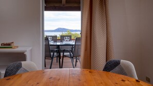 Une table à manger en bois, entourée de chaises, avec une vue sur la mer et les montagnes, visible à travers une porte, et un rideau à motifs.