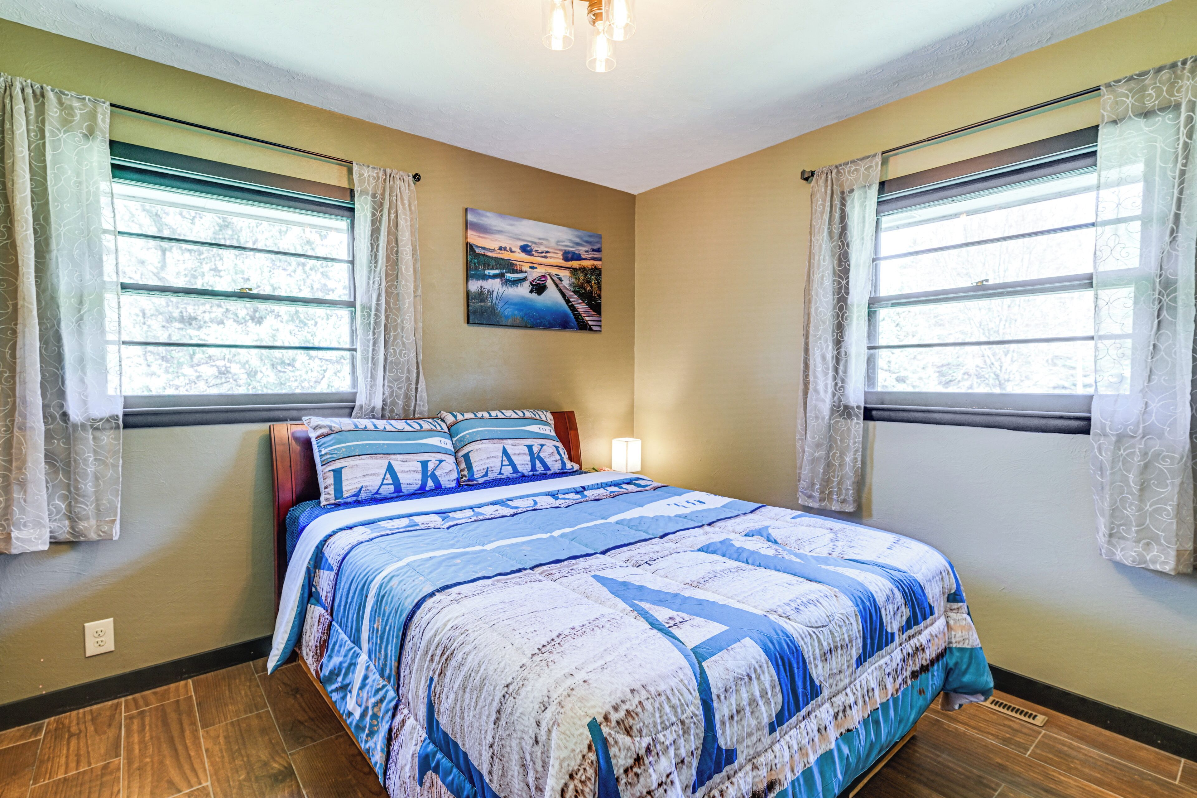 A bedroom with a bed, two windows with curtains, a painting on the wall, and a nightstand with a lamp.