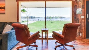 A room with a large window, two leather chairs, a small wooden table with two wine glasses, and a view of a lake.