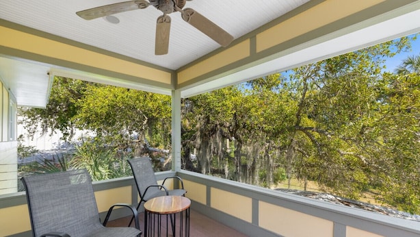 A covered porch with two chairs and a small table, overlooking a natural landscape with trees and a clear sky. - Enjoy the quiet and walk to downtown! (Cedar Key)