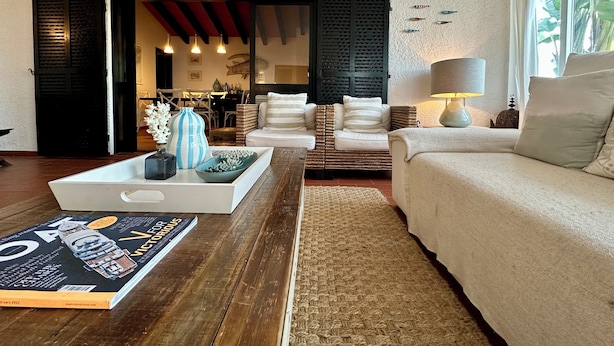 A living room with a sofa, a coffee table with decorative items, a magazine, and a view of the dining area through open doors.