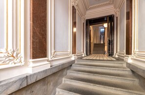 A grand staircase with marble steps and ornate white columns, leading to a well-lit hallway.