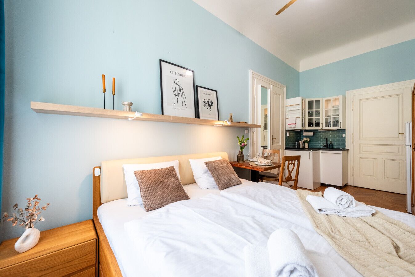 A bedroom with a bed, bedside table, and a small desk with a chair. There is a kitchenette with cabinets and a refrigerator.