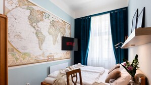 Une chambre d’hôtel bien rangée, avec une grande carte du monde accrochée au mur, un lit recouvert de draps blancs, une table à manger dressée avec des assiettes et des verres, et un téléviseur affichant le logo de Netflix.