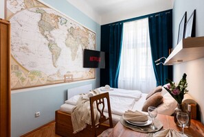 A neatly arranged hotel room with a large world map on the wall, a bed with white linens, a dining table set with plates and glasses, and a television displaying the Netflix logo.