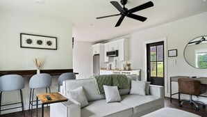 A modern living room with a sofa, a coffee table, and a desk with a chair. There is a ceiling fan, a kitchen area with cabinets, and a window with a view. - 2 Bedroom Beacon Cottage 3 - Two bedrooms, one sleeper sofa and a kitchenette (Allons)