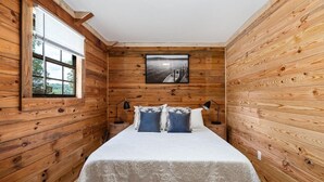 Ein Schlafzimmer mit Holzwänden, einem Bett mit weißer Bettwäsche und zwei Kissen, einem Fenster mit Jalousien und einem Bild an der Wand.