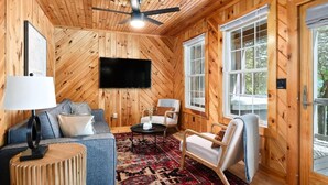 A cozy cabin interior with a wooden ceiling, a ceiling fan, a flat-screen TV mounted on the wall, a sofa with cushions, a chair, a small round table, and a patterned rug. - 10 Person Golden Cedar Lodge 16 - 2 Floors and a 3rd floor loft, entertainment room, full kitchen (Allons)