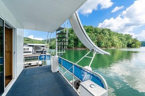 A modern boat deck with a glass railing, a metal staircase, and a view of a calm lake and forested hills. - The Castaway Houseboat 53' - Smallest Houseboat, 3 bedrooms, waterslide, covered rooftop lounge area (Allons)