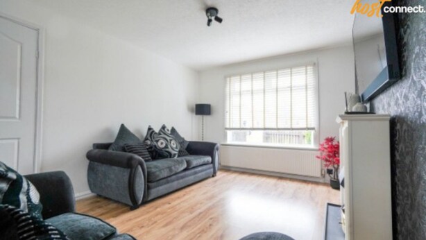 Een ruime woonkamer met een grijze bank, een zwarte fauteuil en een wit kastje.